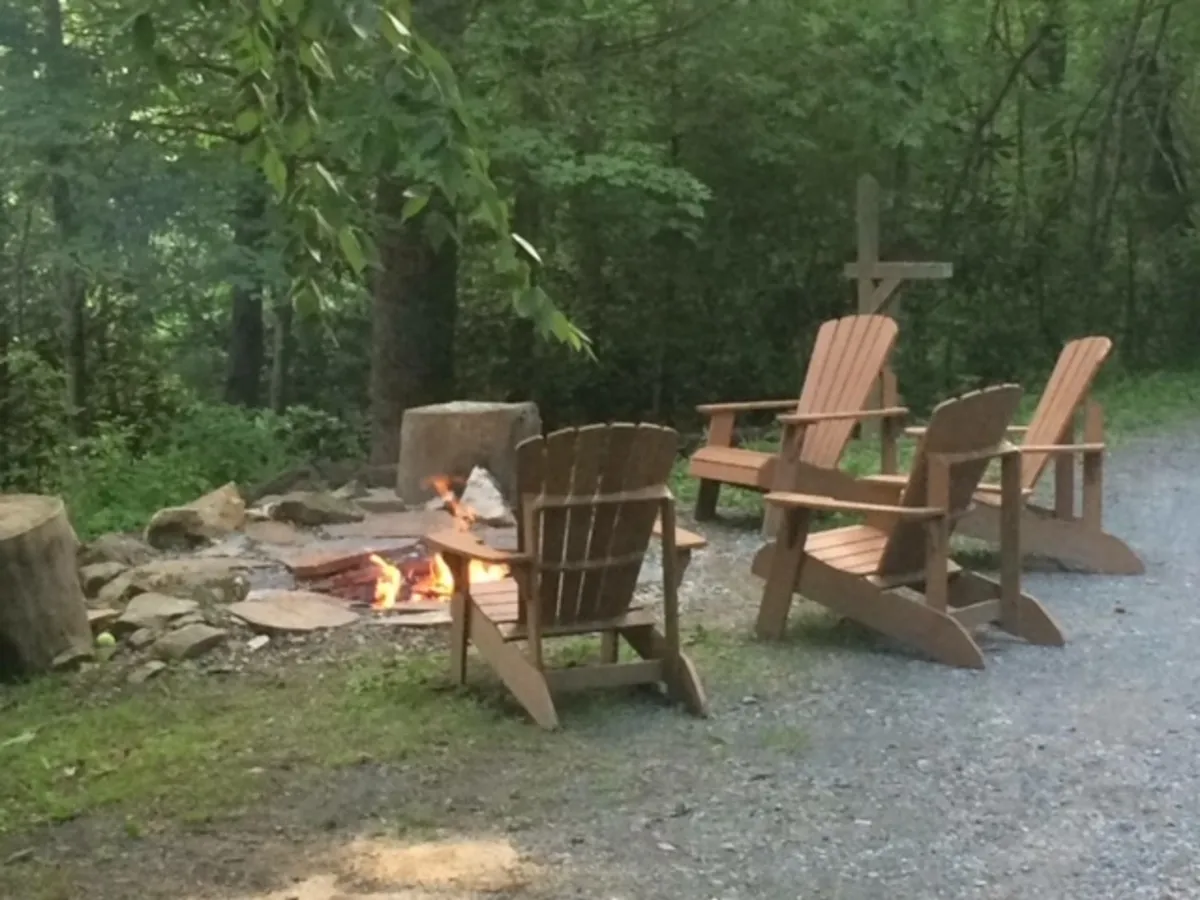 A fire pit encircled by classic Adirondack chairs is nestled among trees, creating a warm, communal space. The area invites guests to gather for storytelling, stargazing, and marshmallow toasts under the stars, embodying the social and cozy aspects of mountain living.
