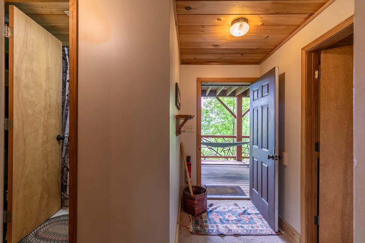 This interior image features an inviting entryway with an open door leading outside, a cozy decorative rug, a wall-mounted shelf perfect for stowing coats or bags, and a glimpse of the natural surroundings beyond the threshold. The wooden ceiling and overall ambiance convey warmth and welcome, while the shelf hints at practical gear storage.