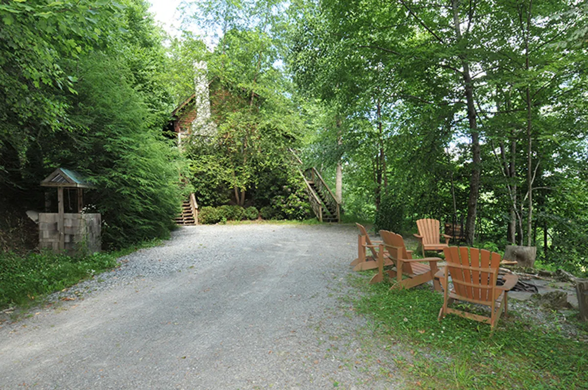 An exterior shot of the cabin shows the gravel driveway leading up to the house, lush greenery surrounding the entrance, and wooden chairs set up for outdoor relaxation. The inviting pathway and fire pit area offer a glimpse of the property’s natural setting and the sense of arrival at this secluded retreat.
