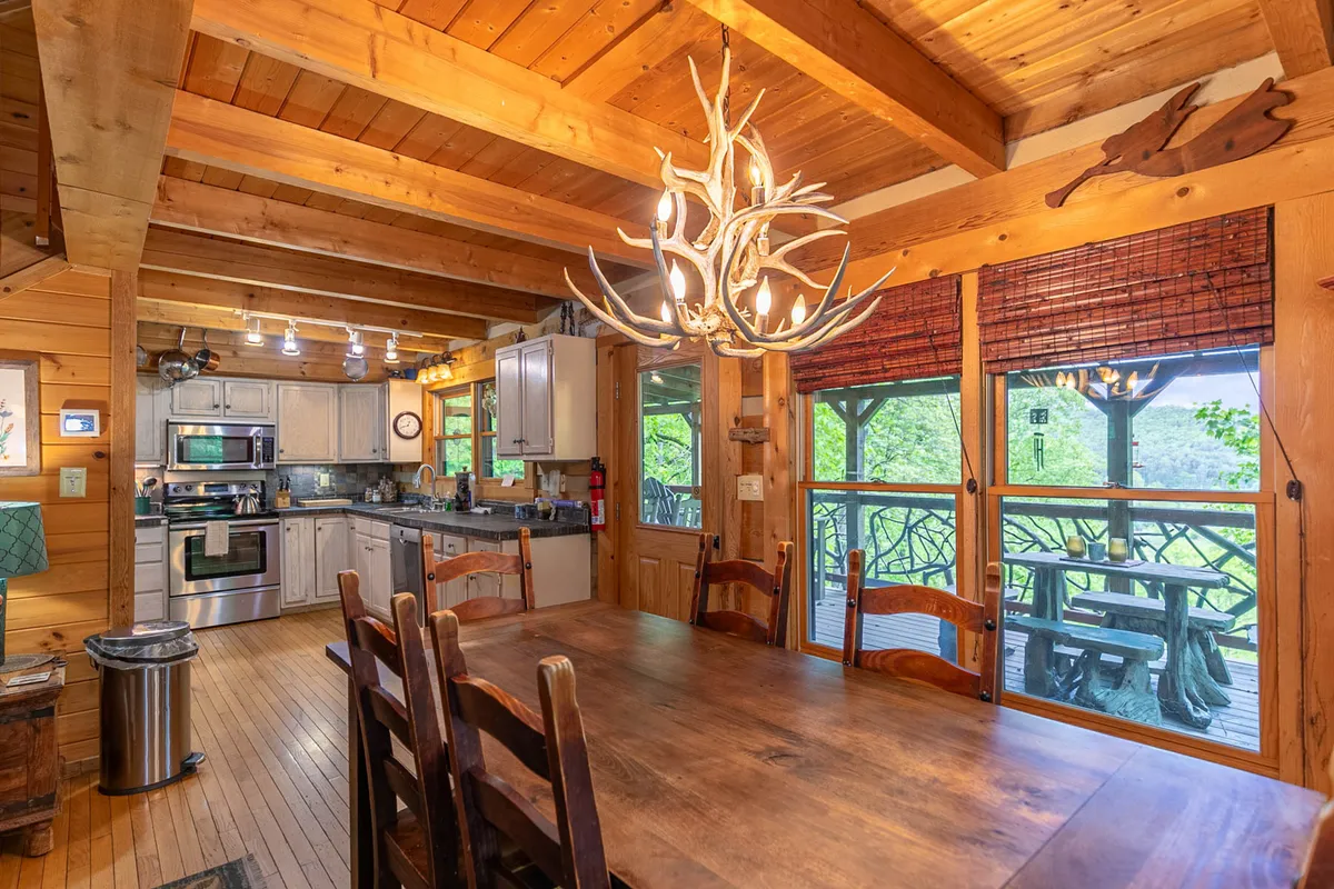 A spacious dining area featuring a large wooden table under an antler chandelier, surrounded by large windows with nature views and glimpses of the open-plan kitchen and modern appliances in the background. The space is set up for shared meals and social gatherings.