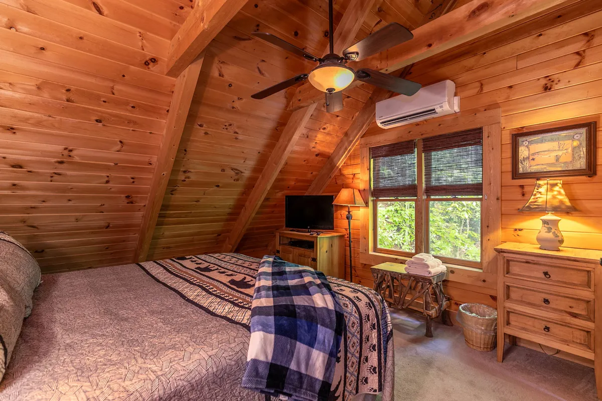 A tranquil bedroom with exposed wooden beams, a comfortable bed with a patterned blanket, a nightstand, and a large window framing lush greenery outside. The space feels serene and connected to nature, perfect for unwinding.