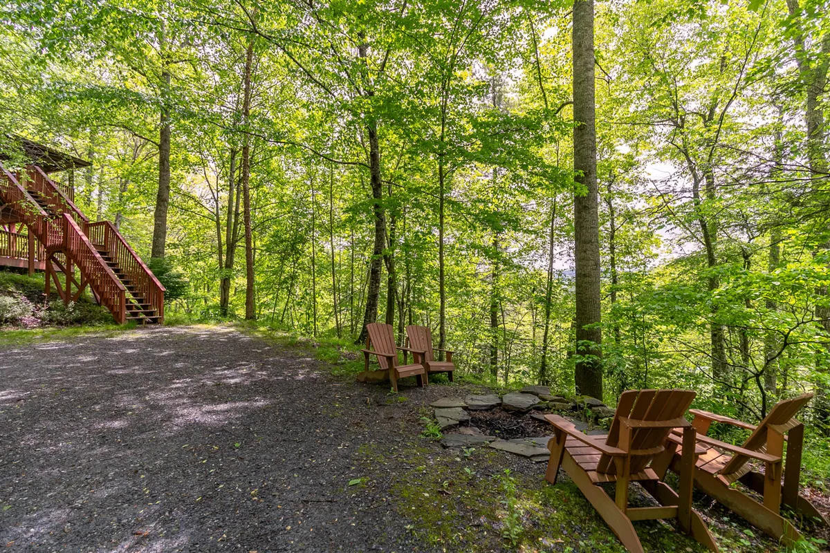 A rustic outdoor space features wooden chairs and a stone fire pit surrounded by trees, with steps leading up to the house. The setting is perfect for relaxing, sharing stories, and stargazing after a day of adventure.