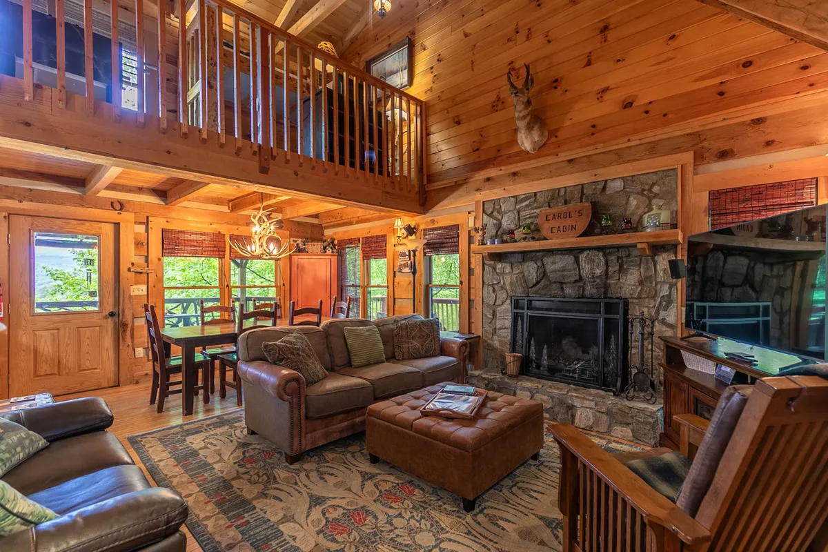 A warm, open living area featuring a stone fireplace surrounded by rustic wooden beams, large windows letting in natural light, and inviting, comfortable furniture. The space is clearly arranged to accommodate family gatherings, with ample seating and a welcoming atmosphere.