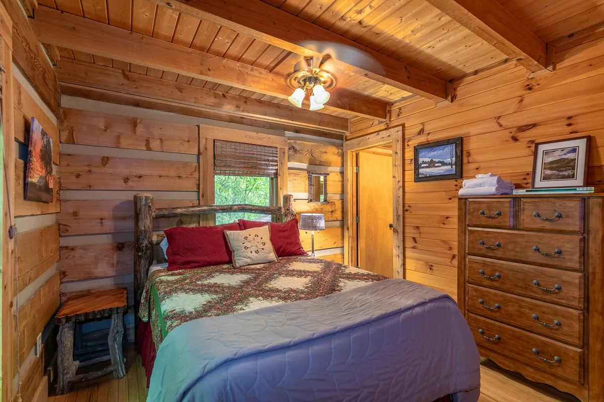 A cozy bedroom featuring a solid wooden bed frame with a colorful quilt, accompanied by a nightstand and lamp. Natural light filters through the window, highlighting the warmth of wooden furnishings and creating a welcoming, restful atmosphere.