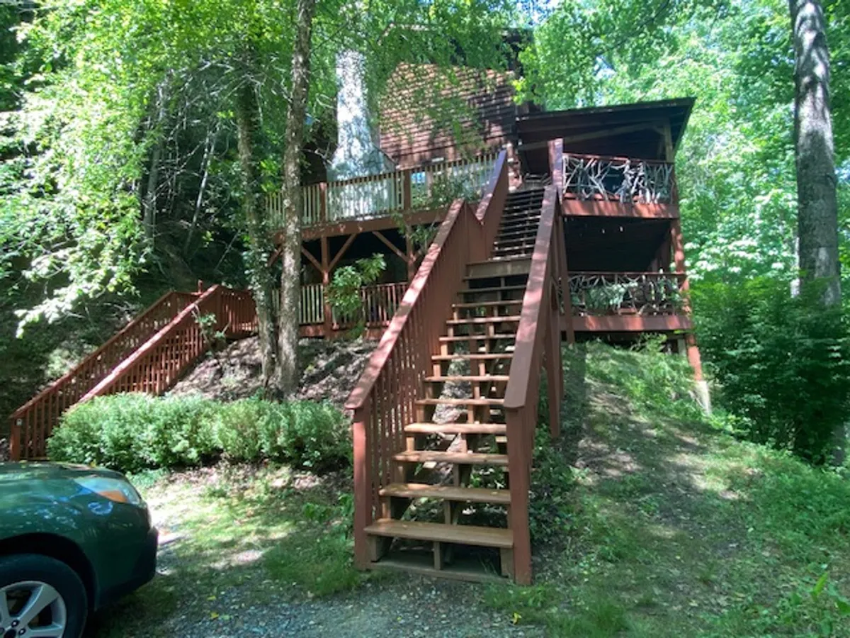 A view of the wooden stairs leading up to a large deck, surrounded by lush greenery. The deck appears inviting for gatherings, with comfortable seating and scenic views beckoning guests outdoors.