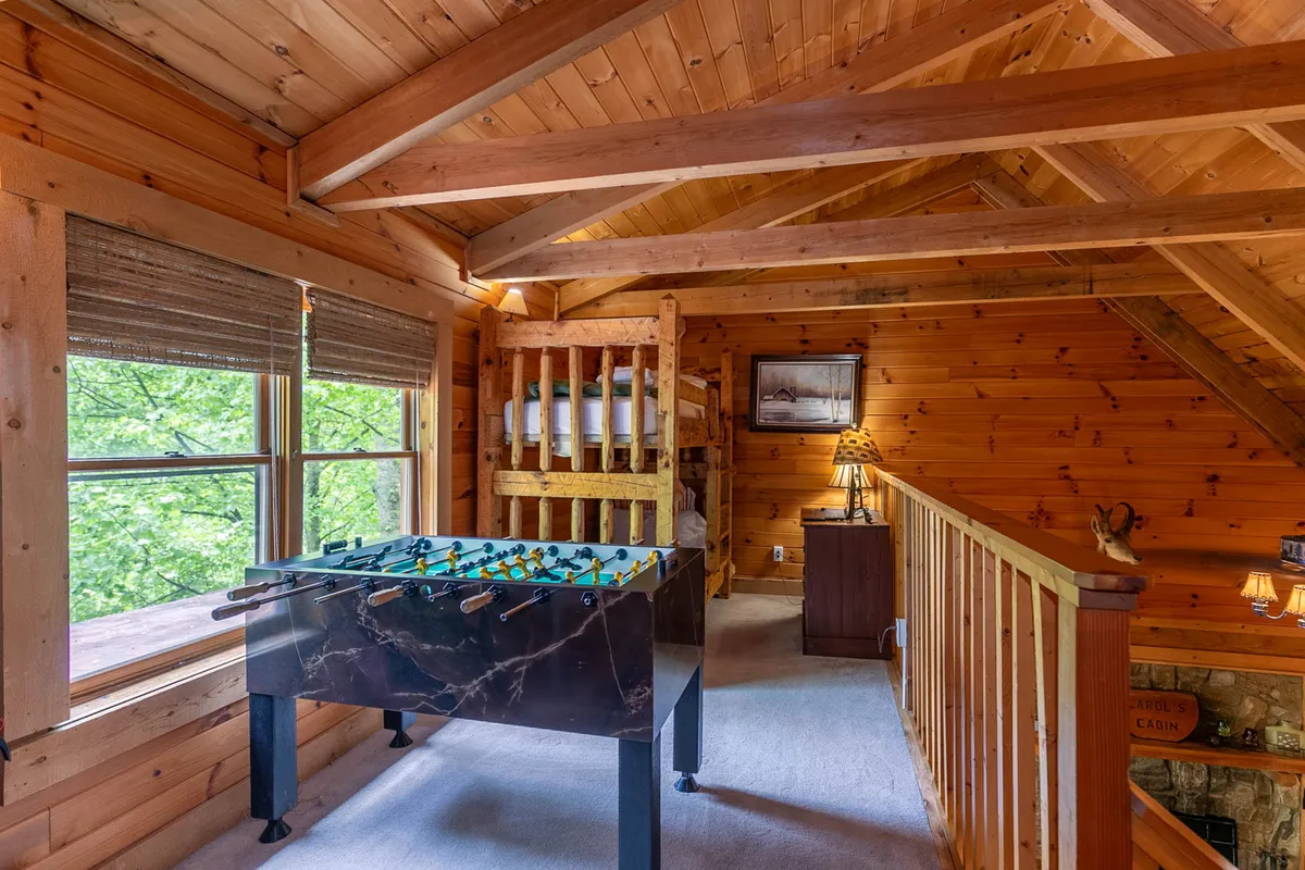 A playful loft space featuring twin bunk beds and a foosball table, accented by warm wood beams, a reading lamp, and a window providing a peek at the outdoors. The room feels youthful and adaptable, perfect for kids or extra guests.