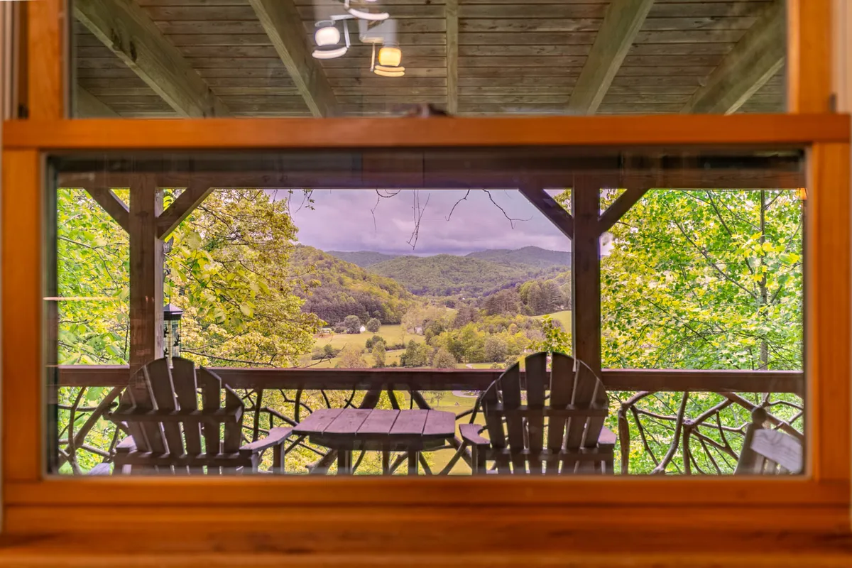 A spacious wooden deck with two Adirondack chairs and a small table, overlooking a stunning mountain vista. The scene exudes tranquility, with a wide-open view perfect for relaxation, reading, or soaking in the natural beauty. The deck is bathed in soft daylight, emphasizing its role as a prime spot for enjoying the Blue Ridge scenery.