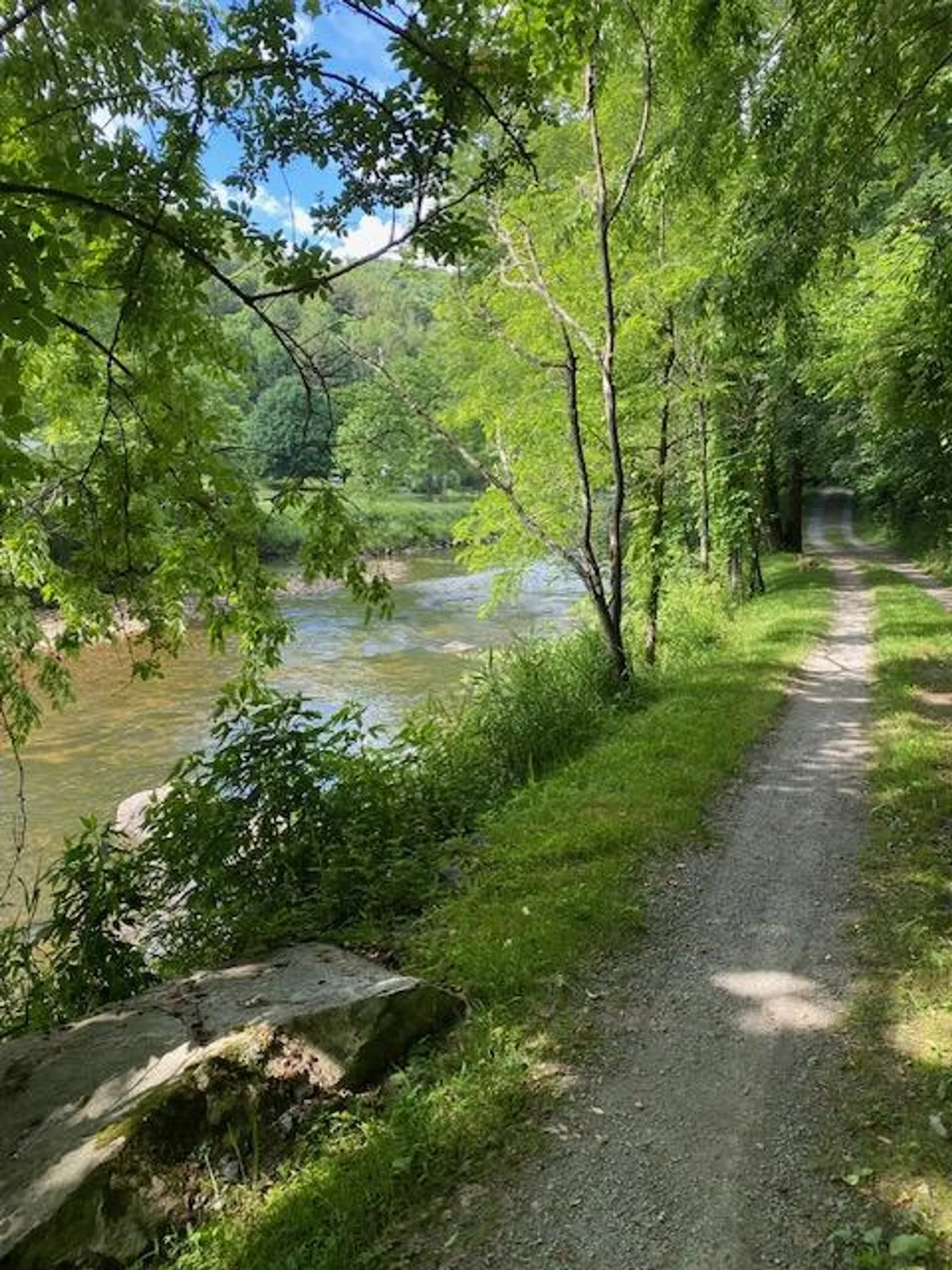 A serene view of a gravel path running alongside a tranquil river, bordered by green foliage and distant trees. This inviting setting suggests a perfect place for walking, biking, fishing, and observing nature.